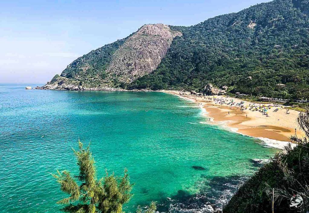 Prainha vista do alto, cercada por densa mata tropical e mar aberto.