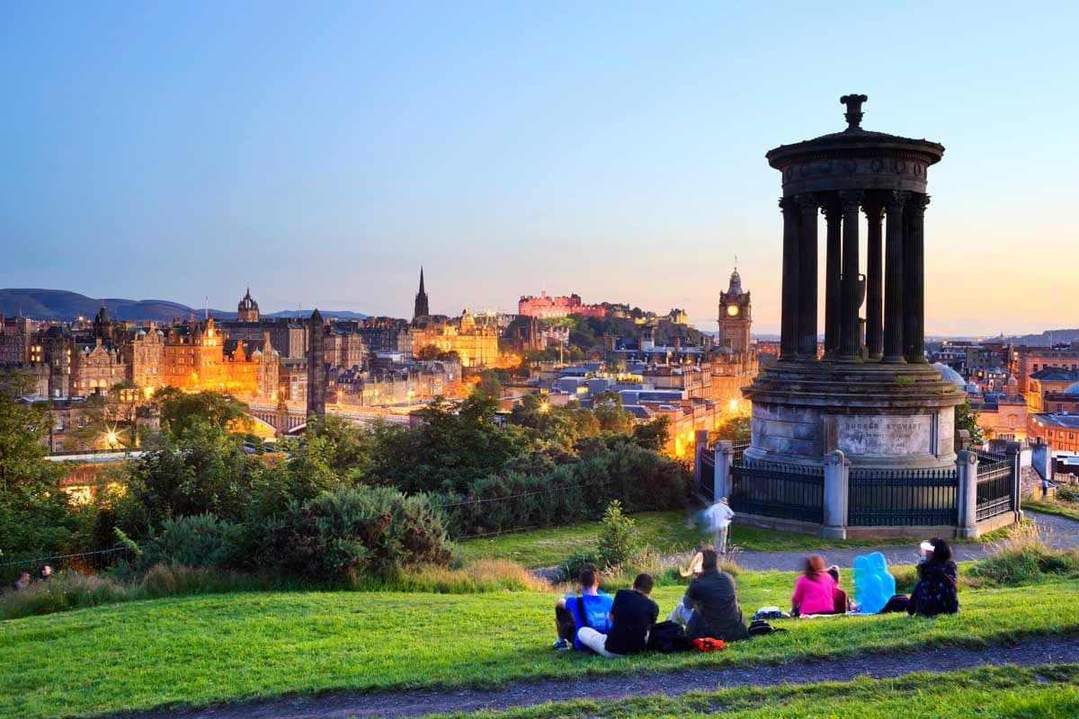 Personas sentados en los pastizales de Calton Hill disfrutando de las vistas panorámicas del casco histórico al atardecer.
