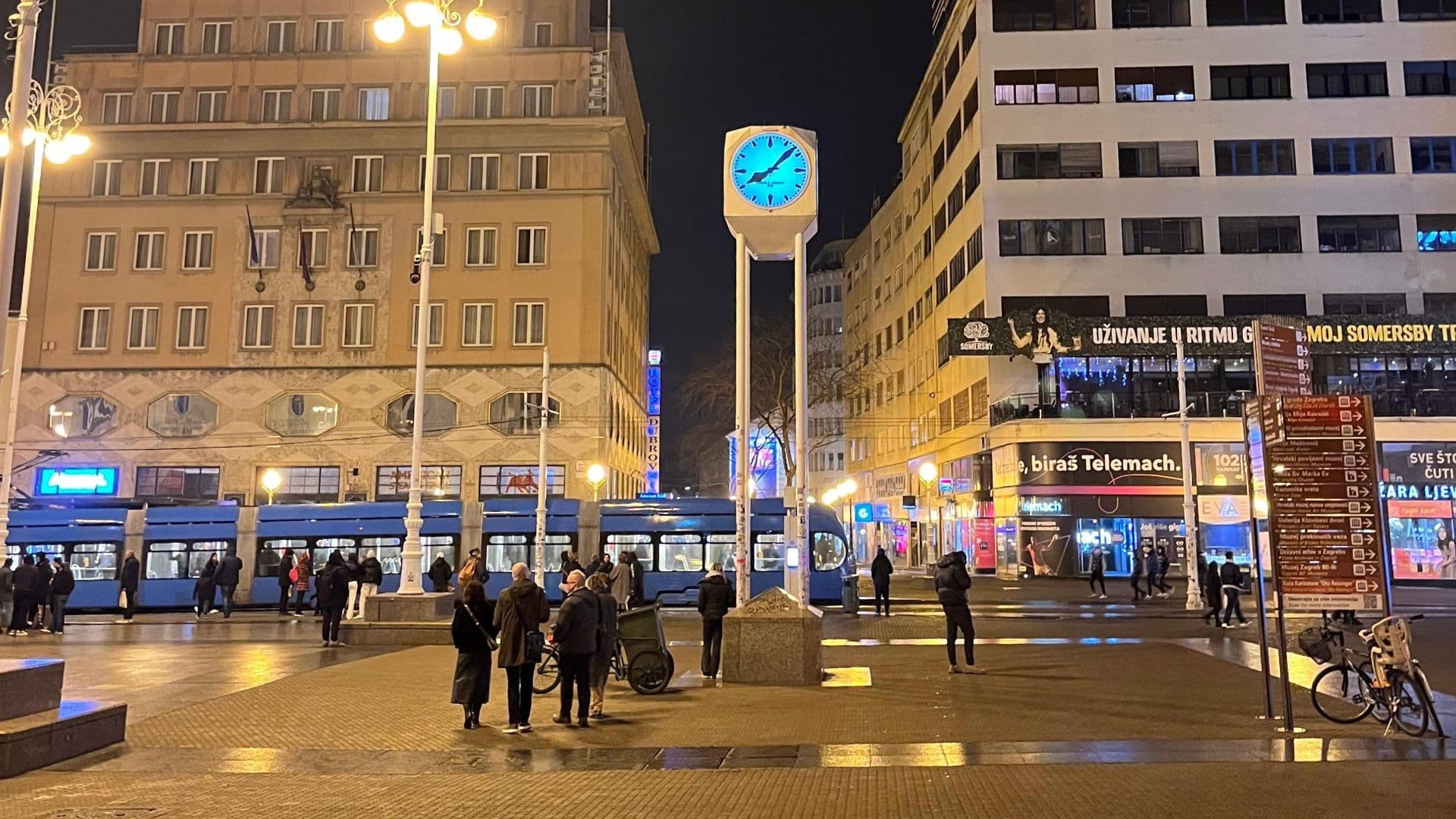 Ban Jelačić Square in Zagreb at night with the blue clock and a tram passing by.