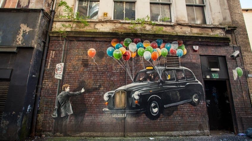 Mural hiperrealista de un taxi negro flotando sostenido por globos de colores en una pared de ladrillo en Glasgow.