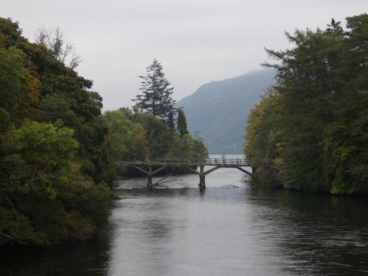 El antiguo puente de madera en Invermoriston. El río Moriston fluye bajo la estructura mientras se abre paso hacia la inmensidad del Lago Ness, que asoma tímidamente al fondo entre la bruma.