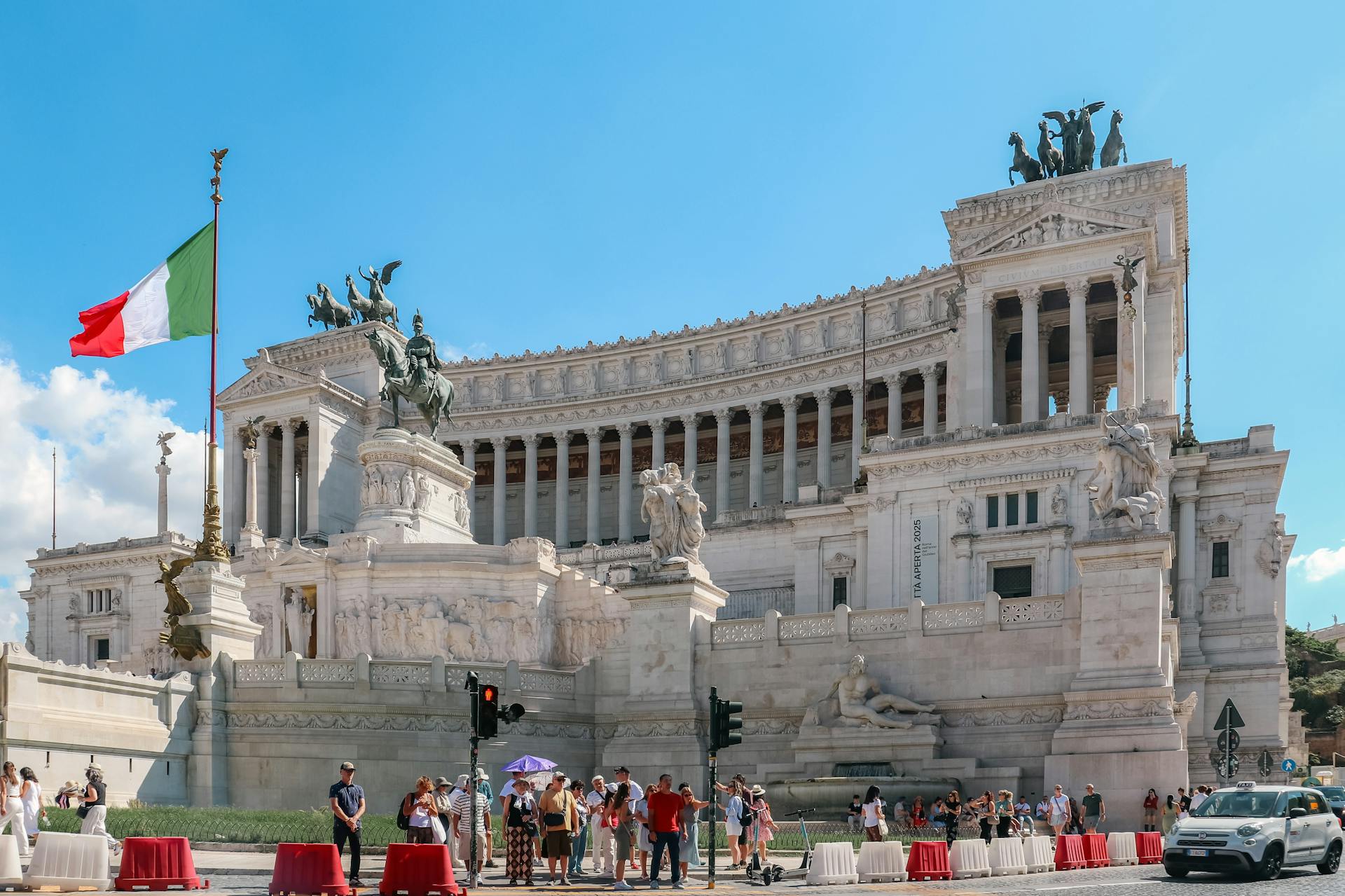 Piazza Venezia. Vista del Altar de la Patria en Roma con visitantes y la bandera italiana ondeando en un día soleado.