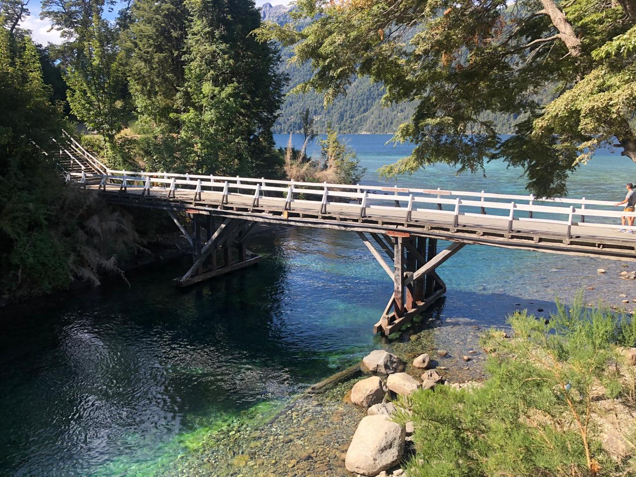 The crystal-clear emerald green water of the Correntoso River passing beneath the pedestrian bridge in Villa La Angostura, Argentina.